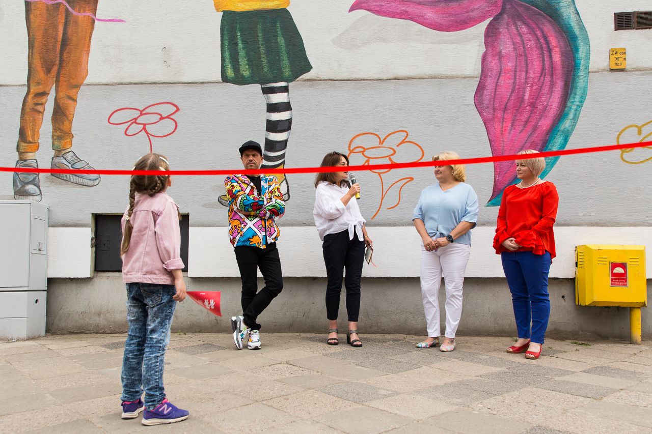 Na chodniku stoi dziewczynka. Spogląda w górę oglądając mural.