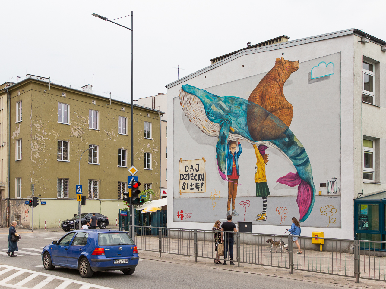 Mural. Dziewczynka i chłopiec trzymają nad głowami wieloryba i niedźwiedzicę. W lewym dolnym rogu muralu napis "daj dziecku siłę!". Na chodniku przechodnie, na jezdni granatowy samochód.