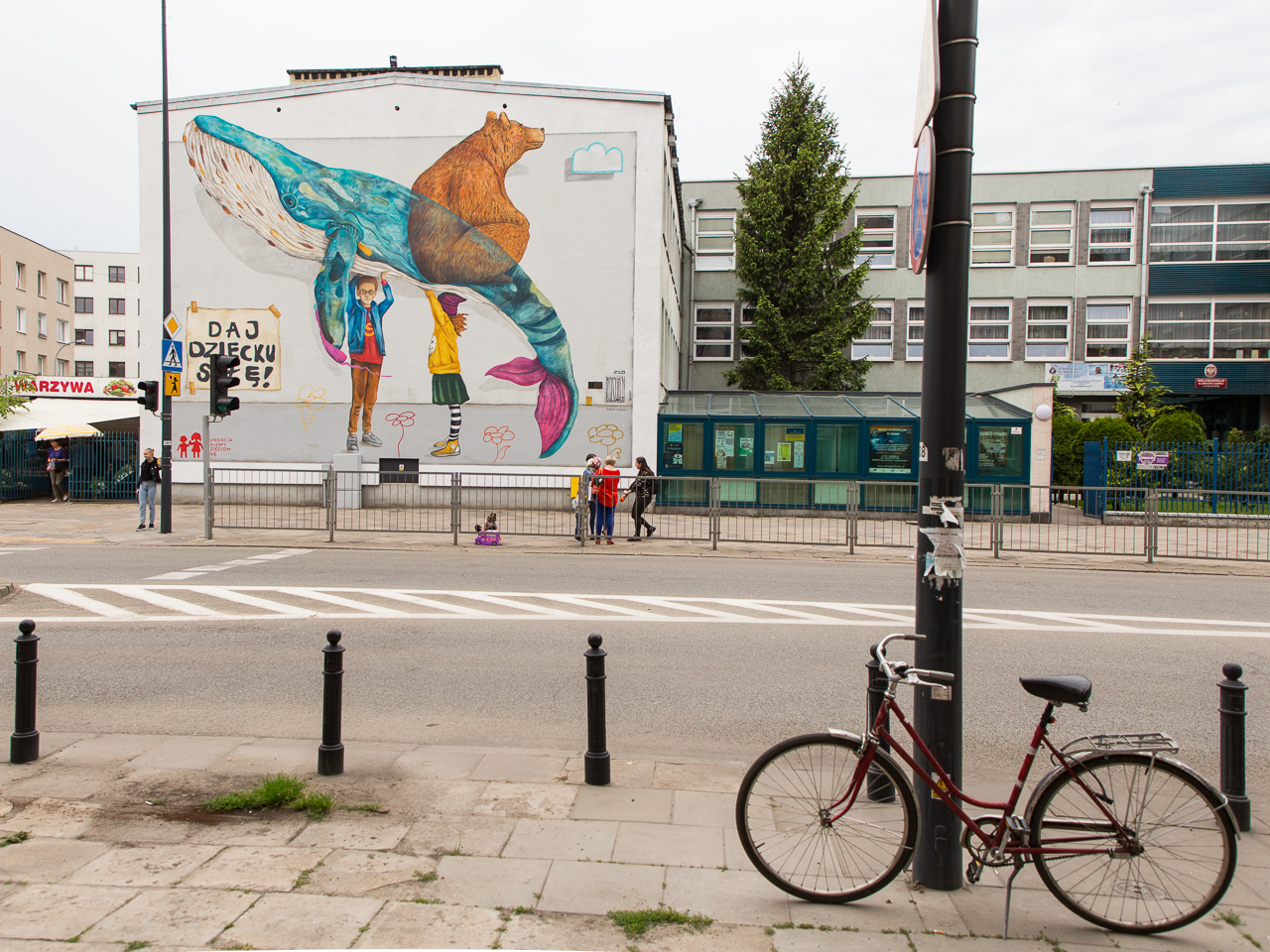 Mural. Dziewczynka i chłopiec trzymają nad głowami wieloryba i niedźwiedzicę. W lewym dolnym rogu muralu napis "daj dziecku siłę!". Na pierwszym planie rower przypięty do słupa.