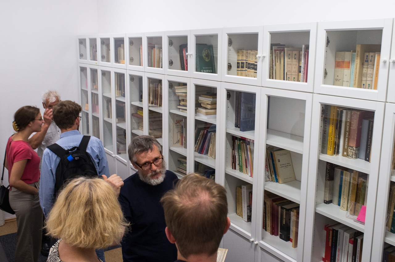 W dużym pomieszczeniu pod ścianą stoją białe szafy biblioteczne. Kilka osób przechadza się, oglądając zawartość szaf. 