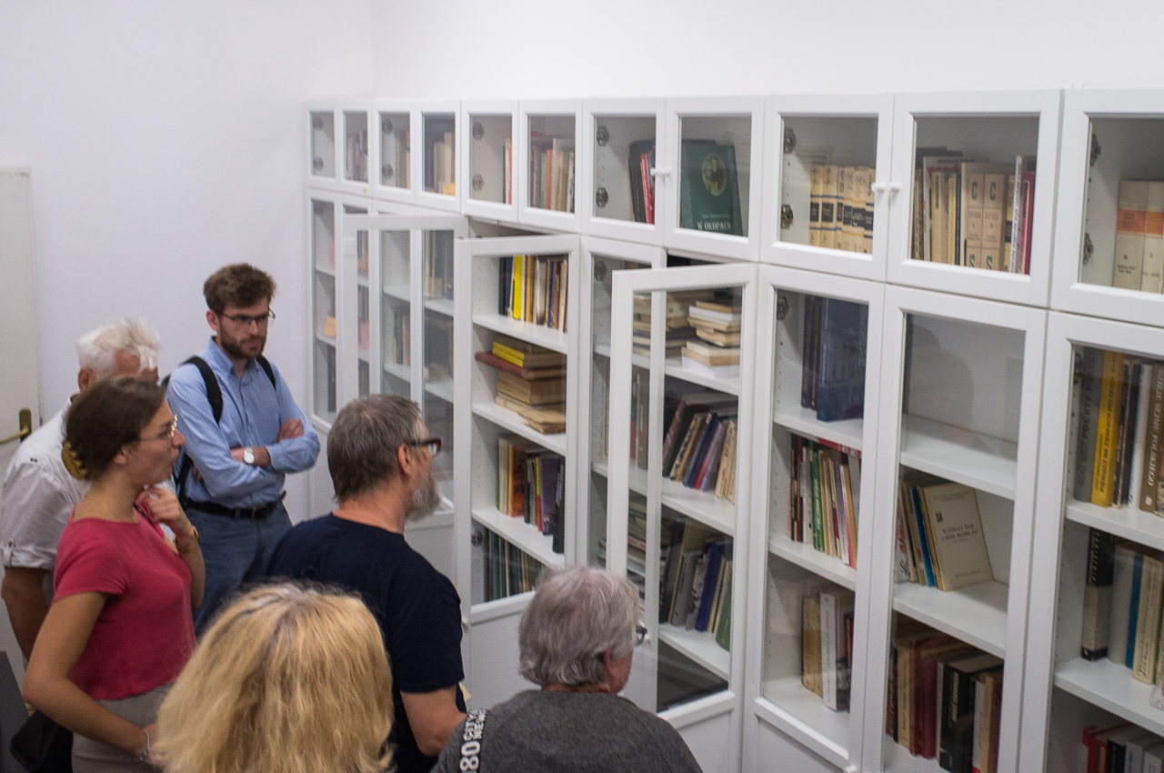 W dużym pomieszczeniu pod ścianą stoją białe szafy biblioteczne. Jedna z nich ma otwarte drzwi. Kilka osób zagląda do środka.