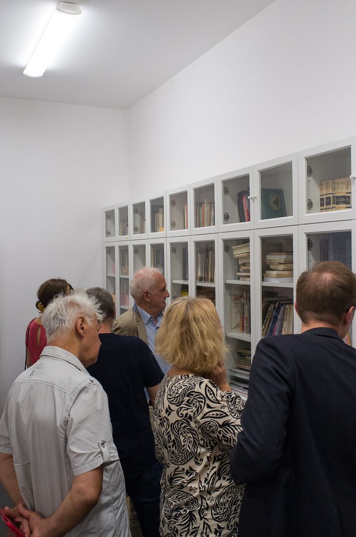 W dużym pomieszczeniu pod ścianą stoją białe szafy biblioteczne. Kilka osób przechadza się, oglądając zawartość szaf. 