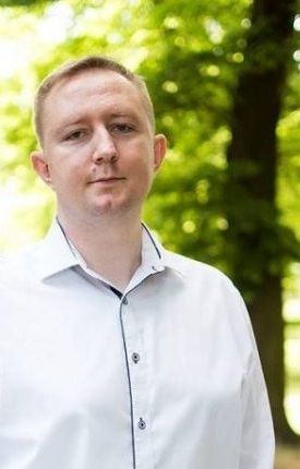 Przemysław Prekiel, Muzeum Historyczne w Przasnyszu. Mężczyzna w białej koszuli stoi na tle zielonych drzew, ze spokojem patrzy w obiektyw, lekko się uśmiecha. Jest po 30-tce, ma włosy koloru blond.