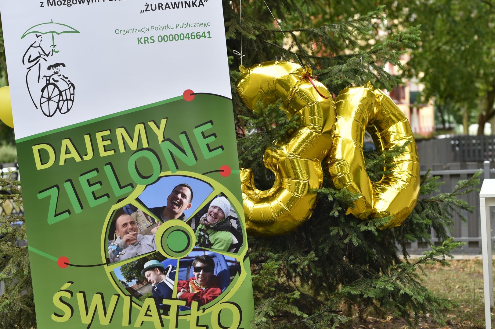 Na zdjęciu widoczny jest banner Stowarzyszenia Żurawinka z napisem "Dajemy zielone światło osobom z MPD" oraz dwa, złote balony tworzące liczbę 30