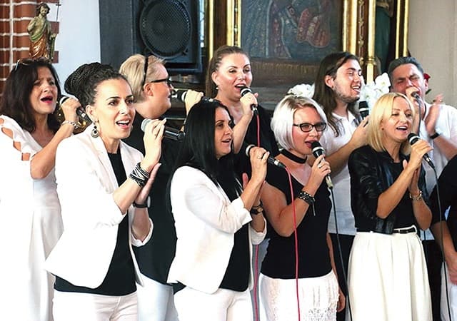 Na zdjęciu widać część chóru - 9 dorosłych osób, stojących w dwóch rzędach: w pierwszym 4 kobiety, w drugim 3 kobiety i 2 mężczyzn. Wszyscy trzymają mikrofony i śpiewają. Ubrani są w biało-czarne stroje.