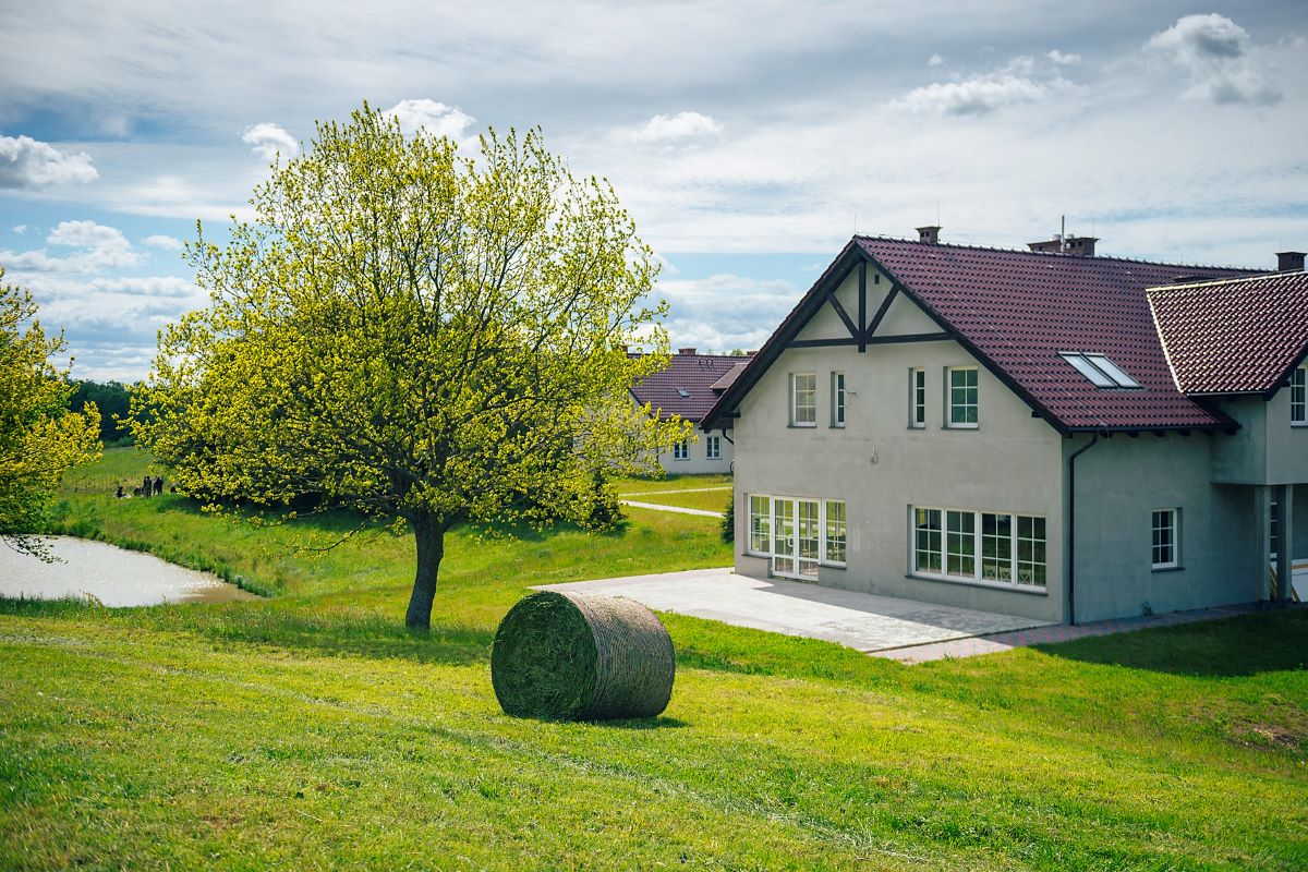 Widok budynku Domu Brzozowego w otoczeniu zieleni łąki i drzewa, z belotem skoszonej trawy