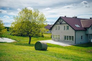 Miniatura Widok budynku Domu Brzozowego w otoczeniu zieleni łąki i drzewa, z belotem skoszonej trawy