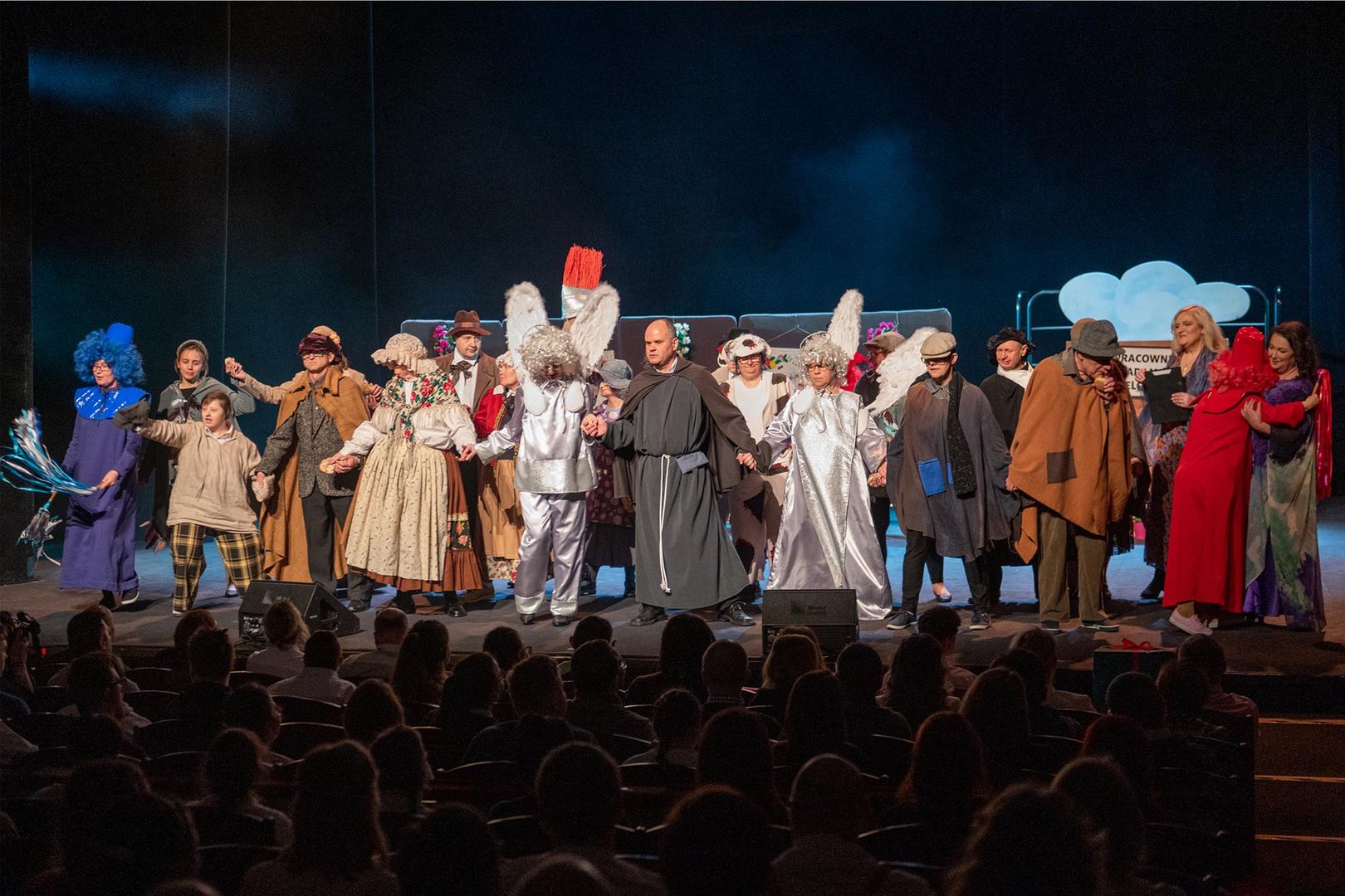 Zdjęcie. Ciemne tło. Grupa aktorów w kostiumach na deskach teatru.