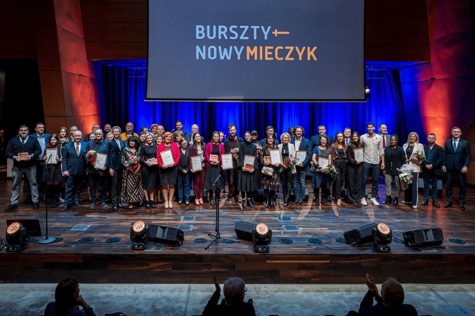 Grupowe zdjęcie laureatów i organizatorów konkursu "Bursztynowy Mieczyk" Na scenie znajduje się duża grupa osób, laureatów konkursu, którzy trzymają dyplomy, kwiaty oraz nagrody. W tle widoczny jest duży ekran z logo konkursu "Bursztyn + Nowy Mieczyk". Uczestnicy pozują do zdjęcia w eleganckiej sali, w otoczeniu niebieskiego i pomarańczowego oświetlenia. W pierwszym rzędzie stoją laureaci z nagrodami, za nimi pozostali goście i organizatorzy.