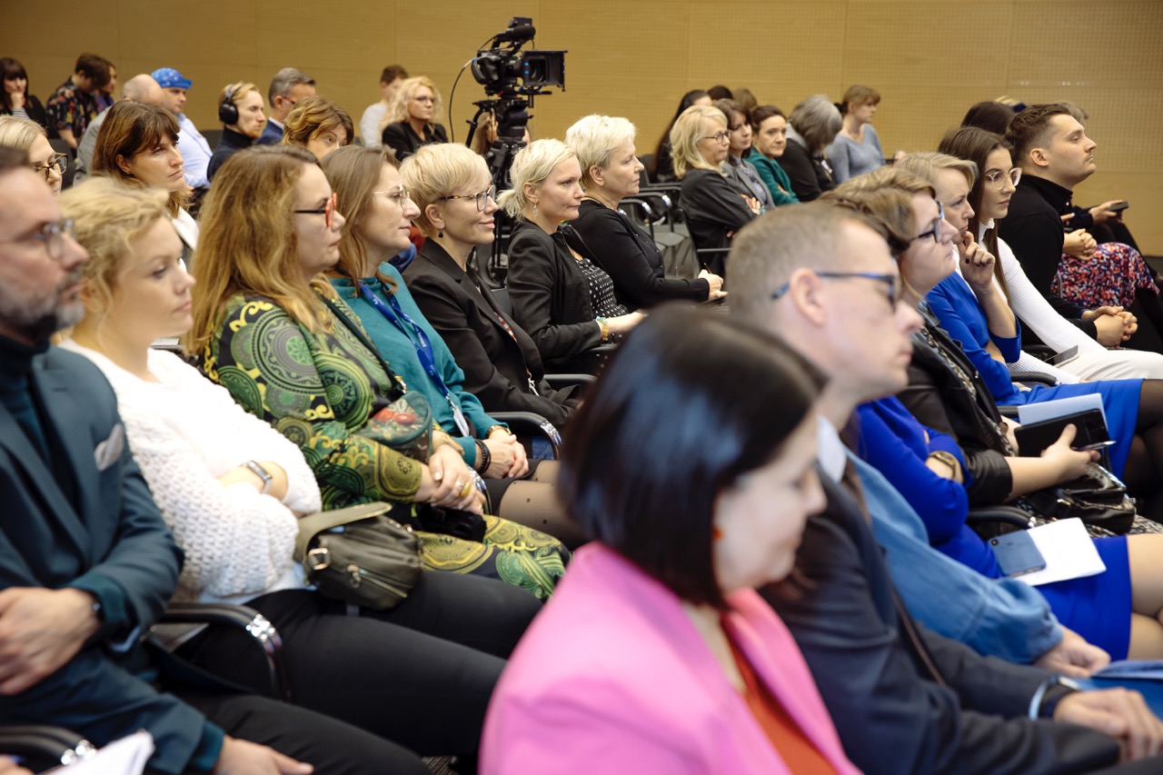 Ujęcie z widowni przedstawiające uczestników siedzących w rzędach podczas czegoś, co przypomina sesję konferencyjną. W tle widać profesjonalną kamerę wideo. Publiczność wydaje się zaangażowana i uważna, a wielu uczestników robi notatki. W pokoju znajdują się drewniane panele ścienne i dobre oświetlenie.