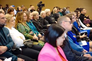 Miniatura Ujęcie z widowni przedstawiające uczestników siedzących w rzędach podczas czegoś, co przypomina sesję konferencyjną. W tle widać profesjonalną kamerę wideo. Publiczność wydaje się zaangażowana i uważna, a wielu uczestników robi notatki. W pokoju znajdują się drewniane panele ścienne i dobre oświetlenie.