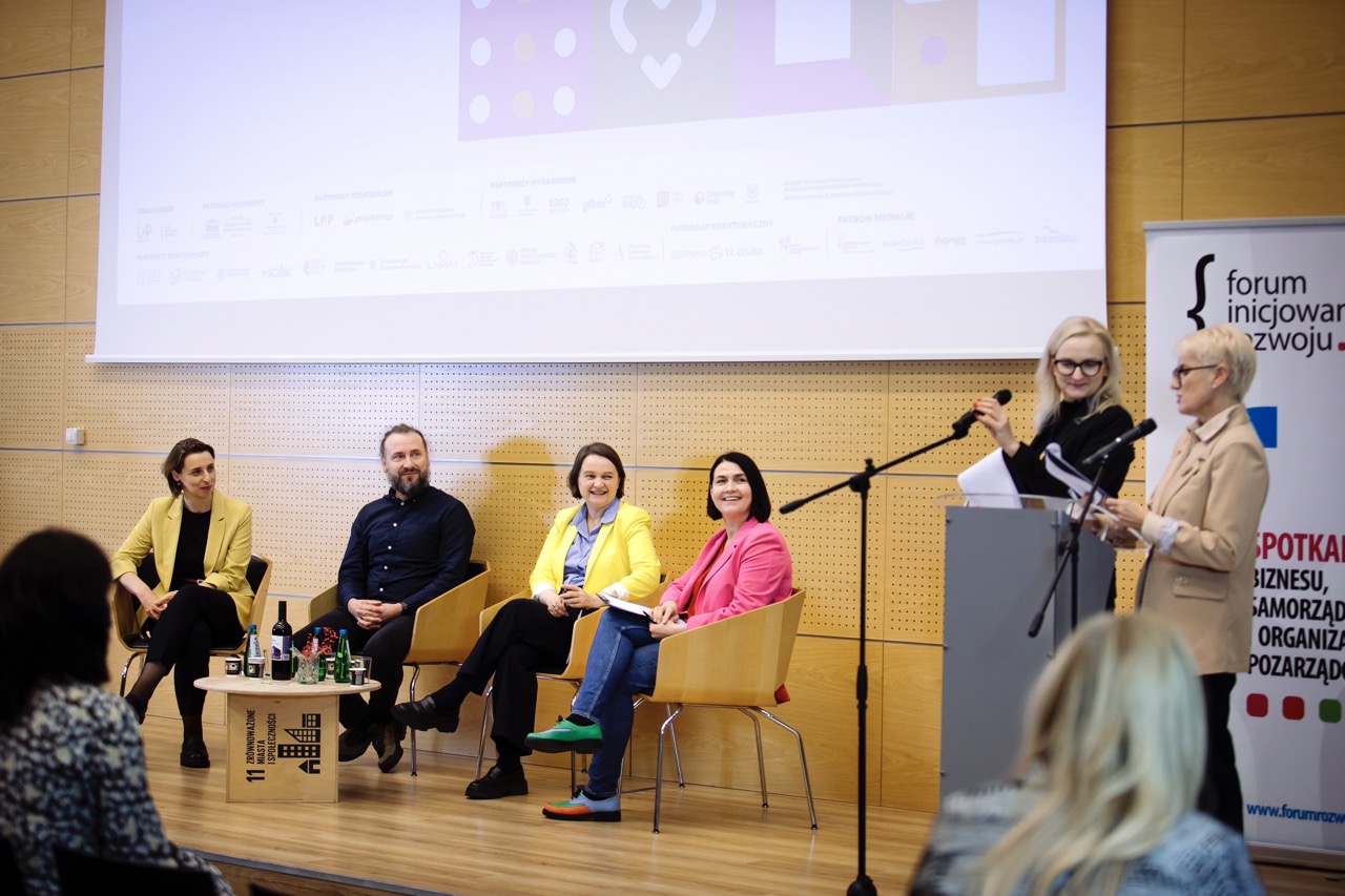 Układ dyskusji panelowej przedstawiający czterech mówców (trzy kobiety i jednego mężczyznę) siedzących na wygodnych krzesłach na scenie. Paneliści są ubrani profesjonalnie, jeden ubrany jest w żółtą kurtkę, drugi w różową. Dwie osoby stoją na podium po prawej stronie. Ustawienie obejmuje ekran prezentacji i baner forum.