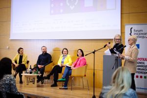 Miniatura Układ dyskusji panelowej przedstawiający czterech mówców (trzy kobiety i jednego mężczyznę) siedzących na wygodnych krzesłach na scenie. Paneliści są ubrani profesjonalnie, jeden ubrany jest w żółtą kurtkę, drugi w różową. Dwie osoby stoją na podium po prawej stronie. Ustawienie obejmuje ekran prezentacji i baner forum.