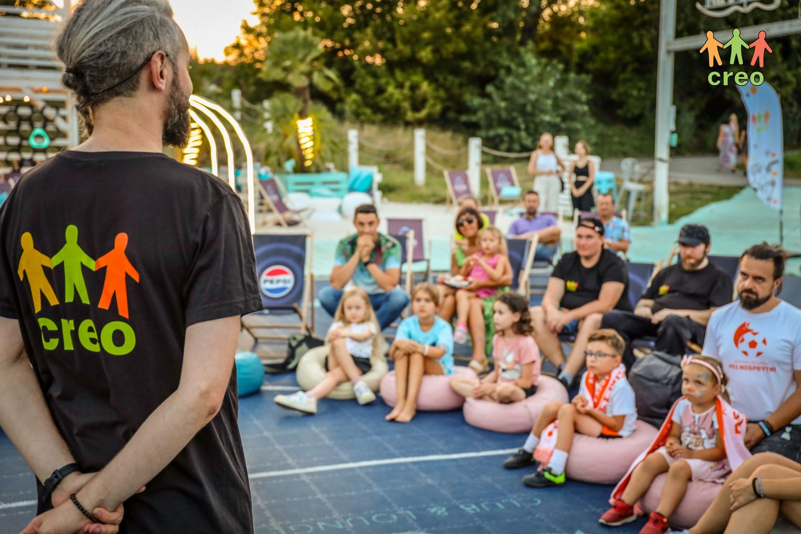 Grupa dzieci i dorosłych siedzi na pufach. Słuchają trenera w czarnej koszulce CREO.