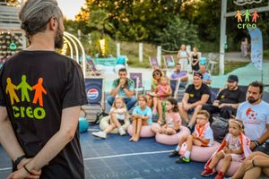 Miniatura Grupa dzieci i dorosłych siedzi na pufach. Słuchają trenera w czarnej koszulce CREO.