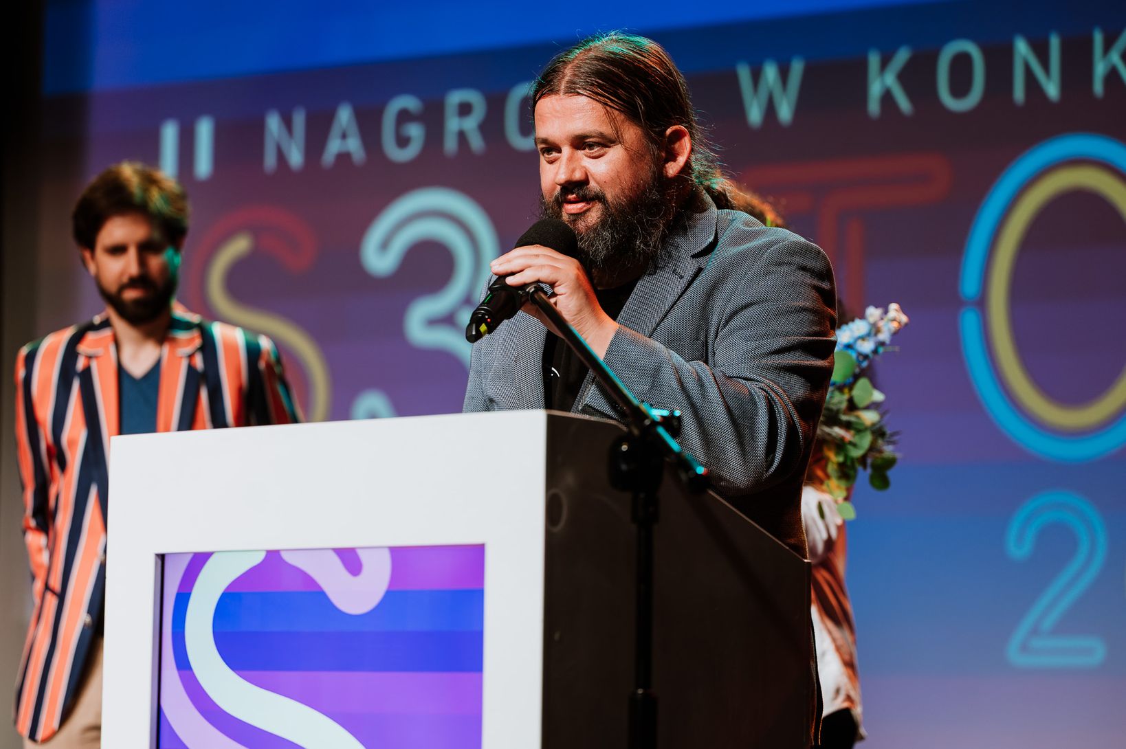 Paweł Zaskalski, mężczyzna z brodą i długimi włosami stoi za podium, przemawiając do mikrofonu. W tle widoczny jest fragment napisu "NAGRODA W KONKURSIE" na dużym ekranie oraz sylwetka drugiego mężczyzny.