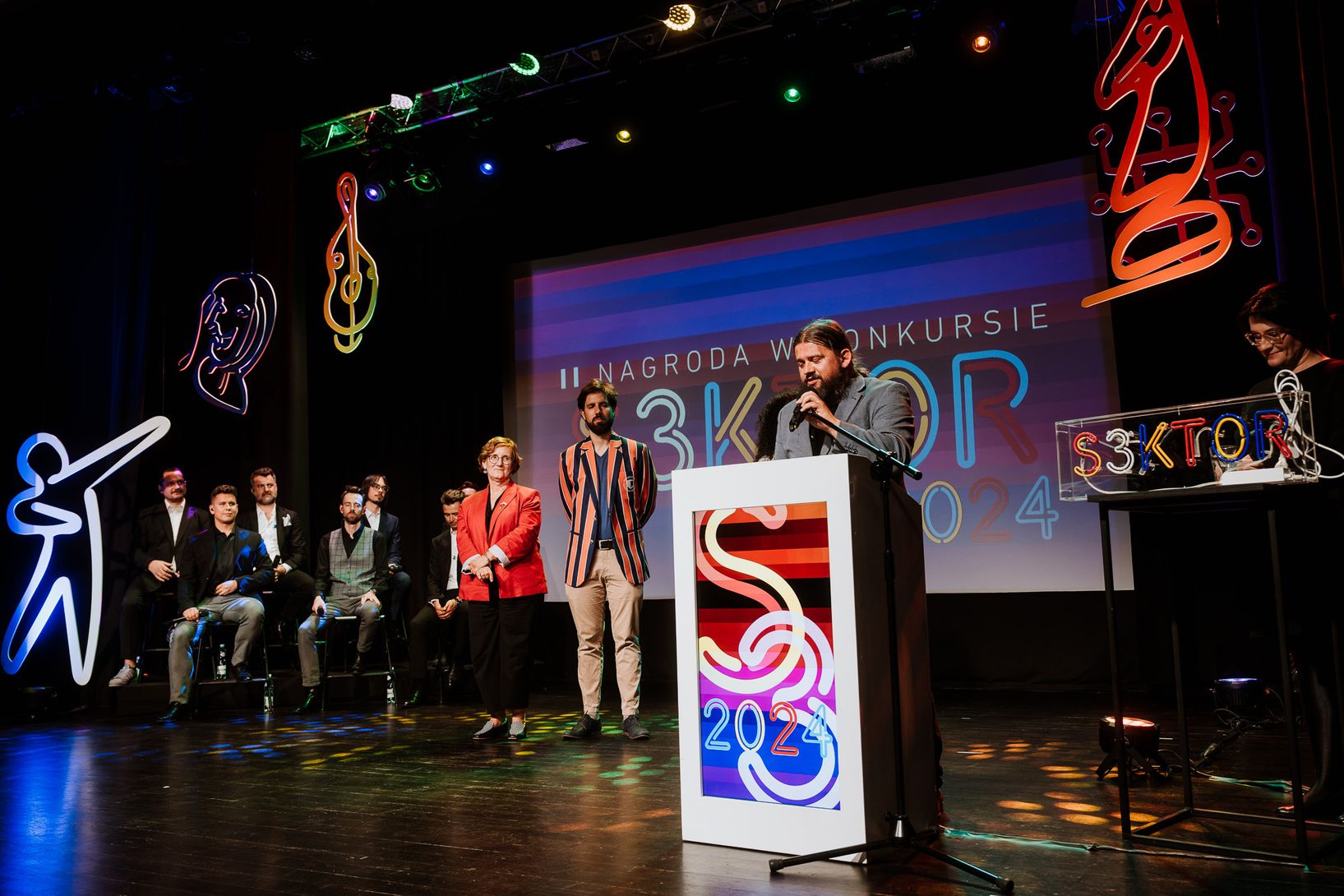 Na scenie, oświetlonej różnobarwnymi reflektorami, mężczyzna z brodą przemawia do mikrofonu za podium. Obok niego stoi kilku innych dorosłych. W tle widoczny jest duży ekran z napisem "NAGRODA W KONKURSIE" i neonowe dekoracje w kształcie figur i liter. Po lewej stronie sceny siedzi grupa mężczyzn, a po prawej kobieta przy konsoli.