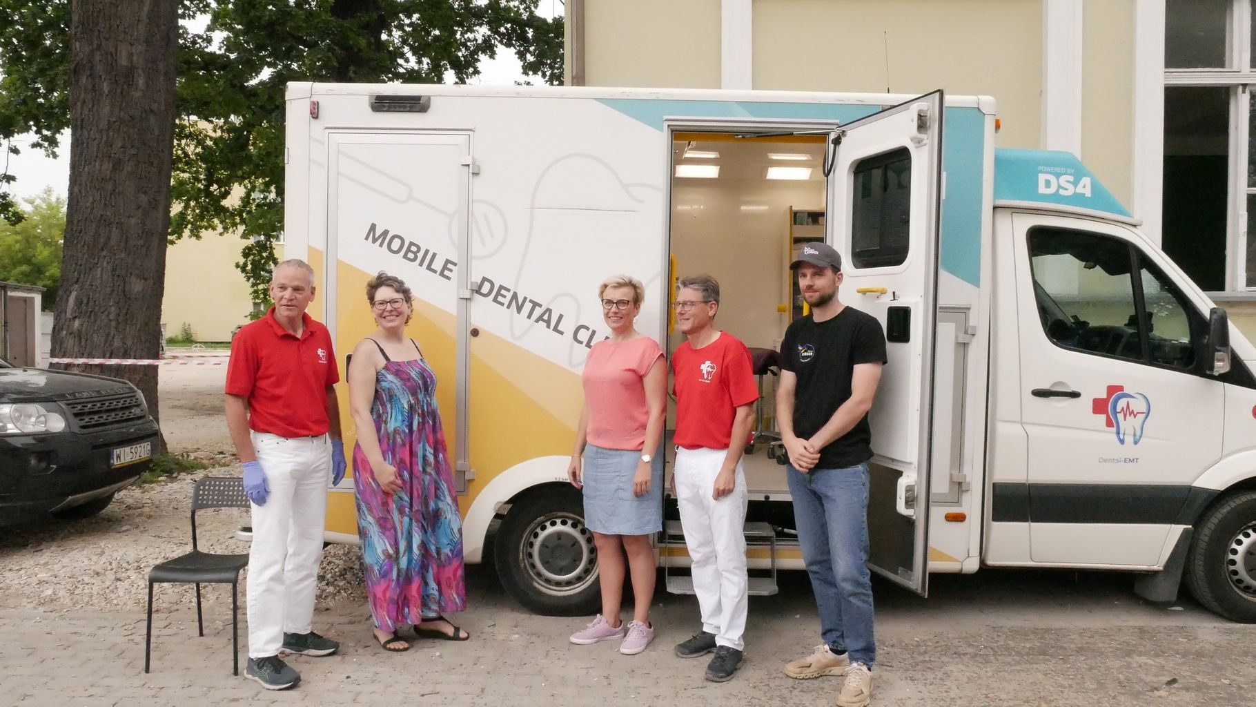  Pięć dorosłych osób stoi przed białym samochodem z napisem "MOBILE DENTAL CLINIC" i otwartymi drzwiami. Po lewej stronie stoi mężczyzna i kobieta, a po prawej kobieta i dwóch mężczyzn. Wszyscy uśmiechają się do obiektywu.