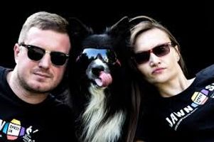 Miniatura Zdjęcie przedstawia dwie osoby w okularach przeciwsłonecznych w koszulkach z napisem "Jawność sprzyja". Między nimi siedzi pies rasy border collie również w okularach przeciwsłonecznych.
