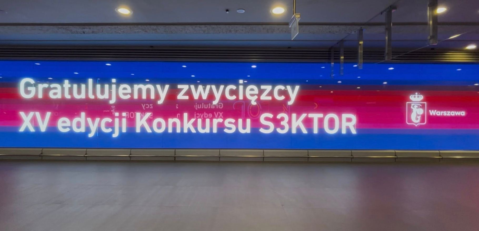  Wielkoformatowy, podświetlany baner na ścianie, z napisem "Gratulujemy zwycięzcy XV edycji Konkursu S3KTOR" oraz herbem Warszawy.