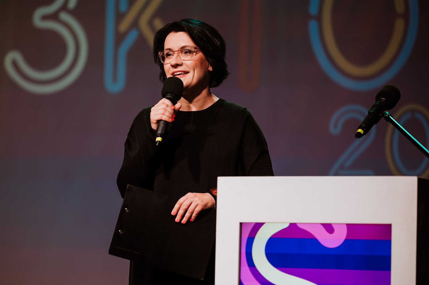  Justyna Dżbik-Kluge.  Kobieta z krótkimi, ciemnymi włosami i okularami, ubrana na czarno, stoi za podium. Uśmiecha się, trzymając mikrofon i czarną teczkę. W tle widoczny jest fragment napisu "S3KTOR".
