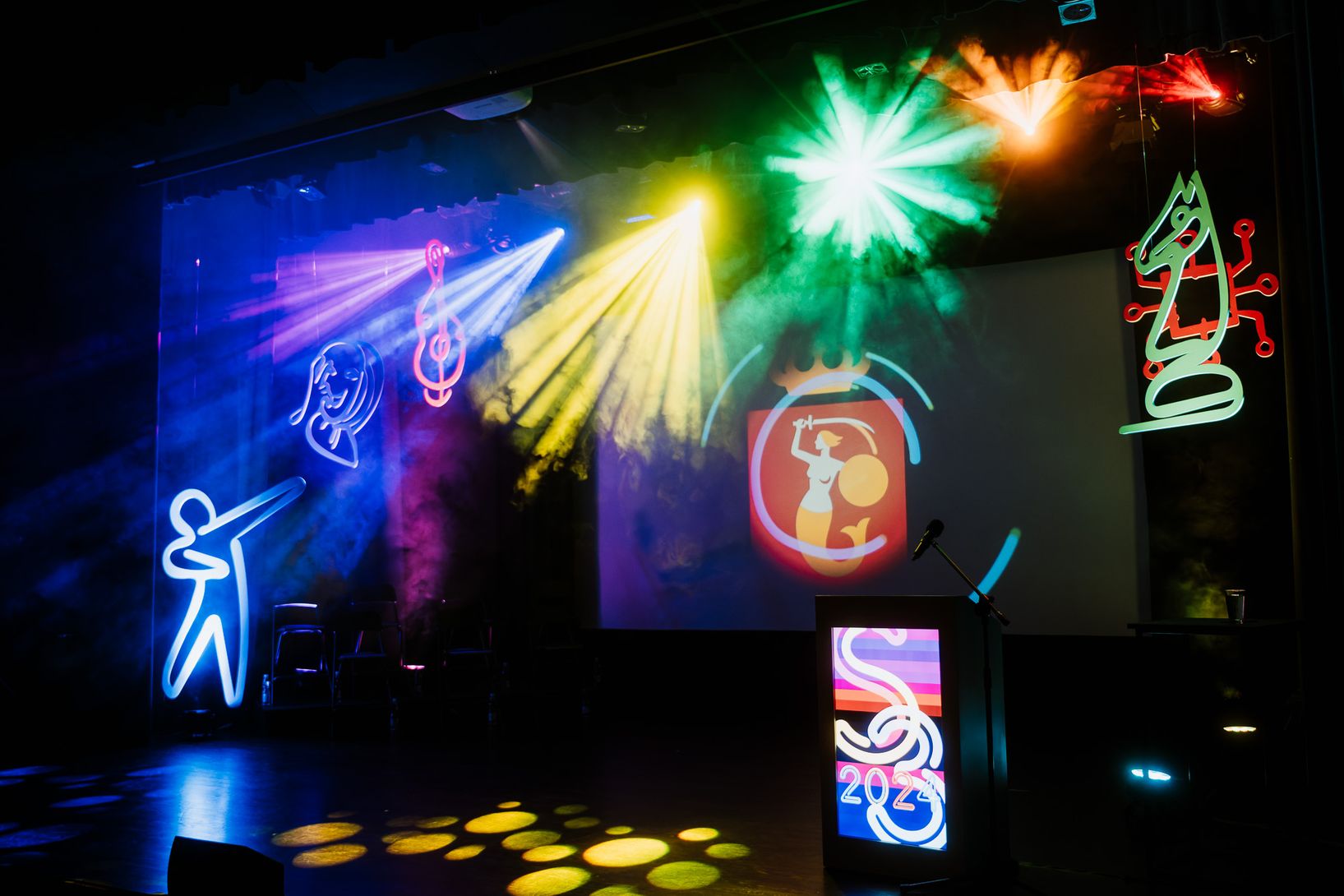 Scena z podium i ekranem, oświetlona kolorowymi reflektorami i neonowymi dekoracjami w kształcie postaci i instrumentów. Na ekranie widoczne jest logo herbu Warszawy.