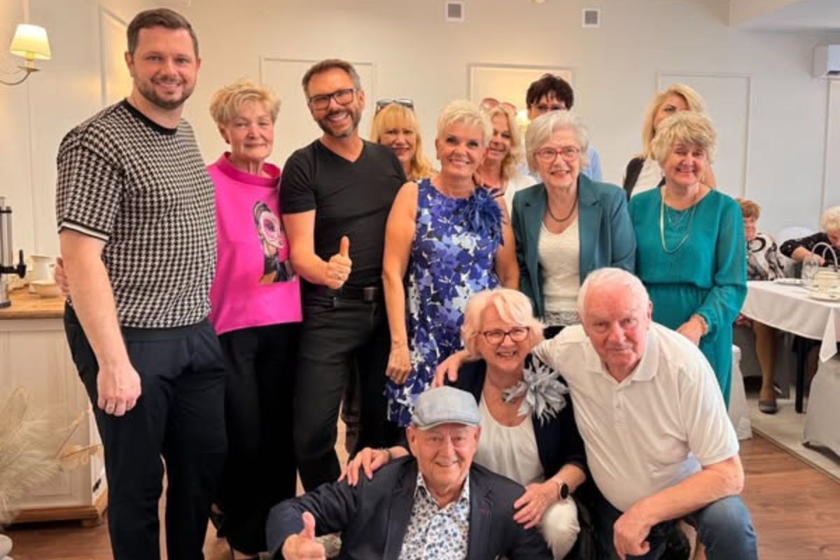  Grupowe zdjęcie przedstawiające kilkanaście uśmiechniętych osób, w większości w wieku senioralnym, w eleganckim wnętrzu, prawdopodobnie restauracji lub kawiarni.  Osoby stoją i kucają, tworząc zwartą grupę. Wśród nich jest młodszy mężczyzna w czarno-białej, wzorzystej koszulce i ciemnych spodniach, uśmiechający się do obiektywu, oraz inny, równie młody mężczyzna, w czarnej koszulce i spodniach, robiący gest kciukiem w górę. Kobiety w grupie mają różnorodne stroje, w tym jaskrawo różowy top, niebieską kwiecistą sukienkę i elegancki turkusowy żakiet. Osoby na pierwszym planie to starszy mężczyzna w czapce z daszkiem i marynarce, kucający i pokazujący kciuk w górę, oraz kobieta i starszy mężczyzna, którzy obejmują się ramionami. Tło jest jasne, widać zasłony i elementy wystroju wnętrza.