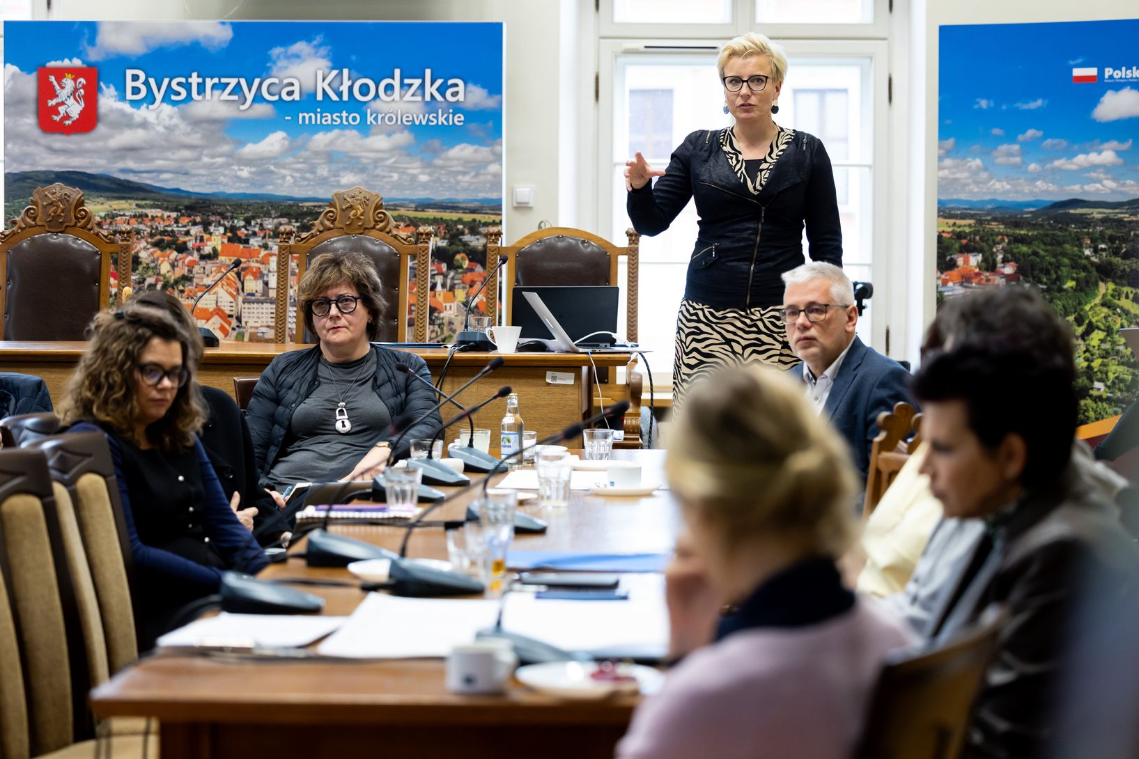 Kobieta o jasnych włosach w okularach, ubrana w czarną, zapinaną kurtkę i spódnicę w zebrę, stoi, przemawiając do grupy osób siedzących wokół stołu. Kobieta gestykuluje dłonią. Na tle za nią widoczne są duże plakaty z nazwą "Bystrzyca Kłodzka - miasto królewskie" i elementami graficznymi.