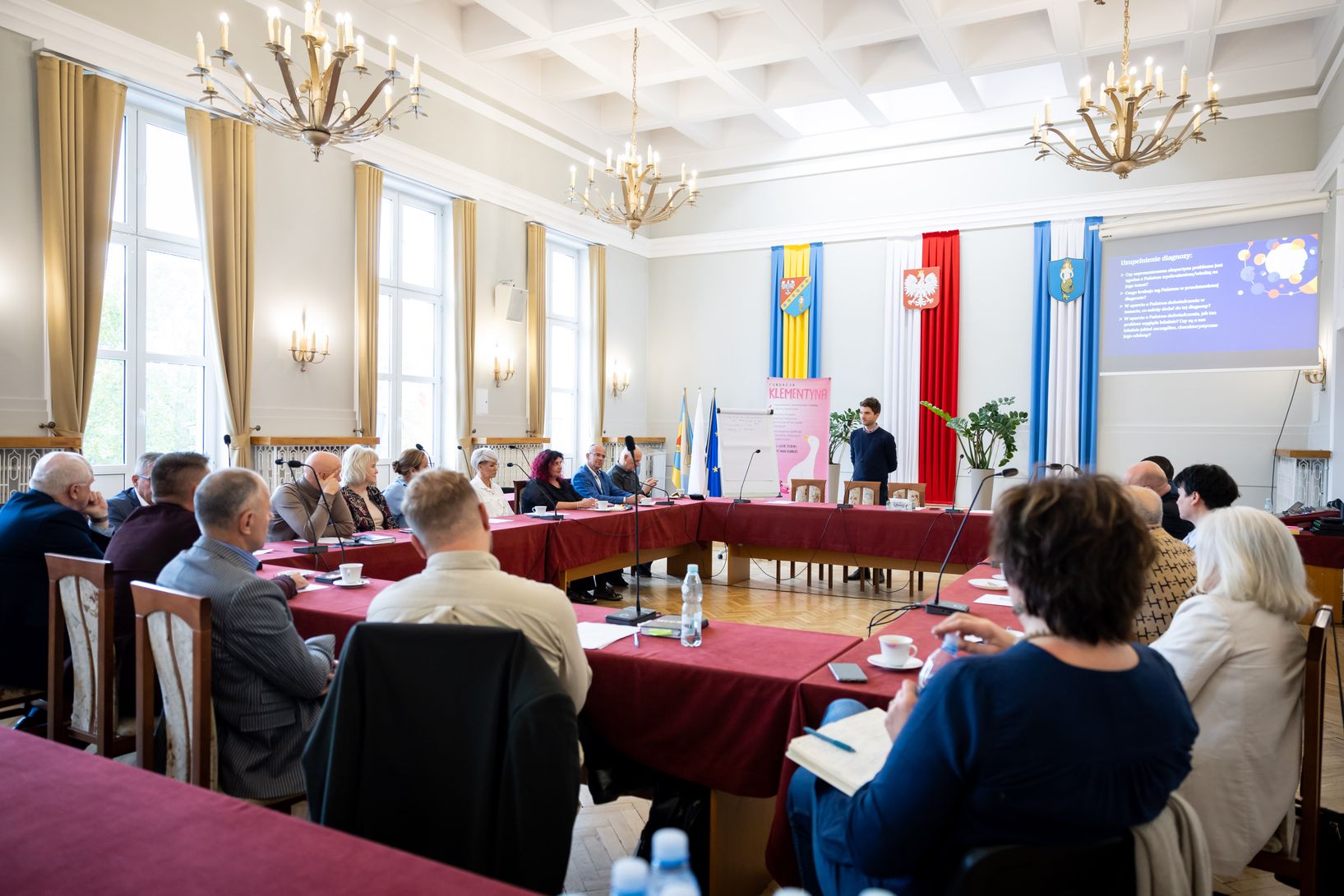Grupa około dwudziestu osób siedzi przy dużym, prostokątnym stole nakrytym bordowym obrusem, w eleganckiej sali. Pomieszczenie ma wysokie okna i trzy duże żyrandole. Na środku stołu, w głębi, stoi młody mężczyzna, przemawiający do zebranych. W tle wiszą flagi, w tym polska, a na ekranie widoczna jest prezentacja.