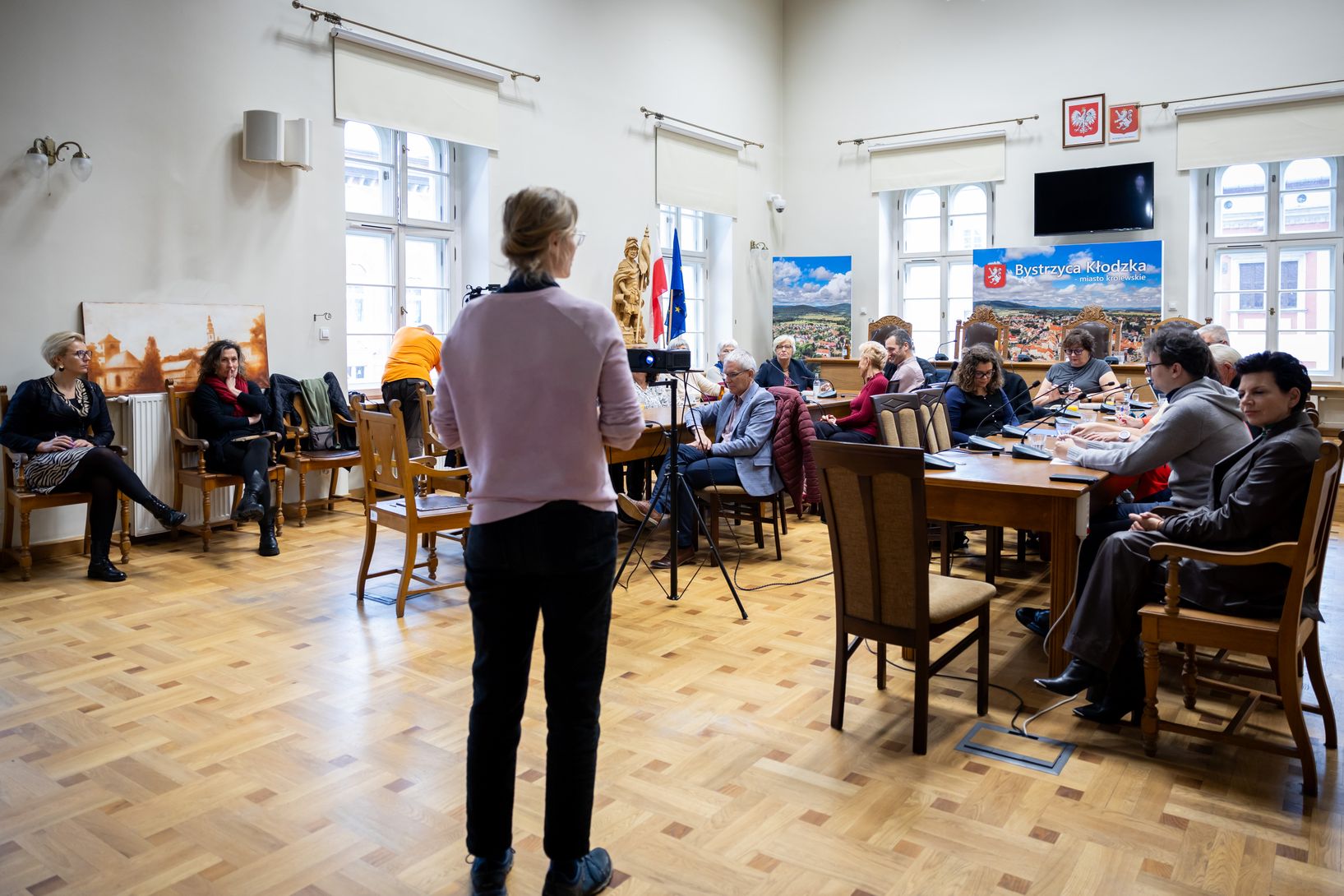 Kobieta o jasnych włosach, w jasnoróżowym swetrze i ciemnych spodniach, stoi tyłem do kamery na środku przestronnej, jasnej sali z drewnianą podłogą w jodełkę. W tle, przy prawym brzegu kadru, grupa osób siedzi wokół drewnianego stołu, uczestnicząc w naradzie. W pomieszczeniu widoczne są duże okna, telewizor oraz plakaty promujące "Bystrzycę Kłodzką".