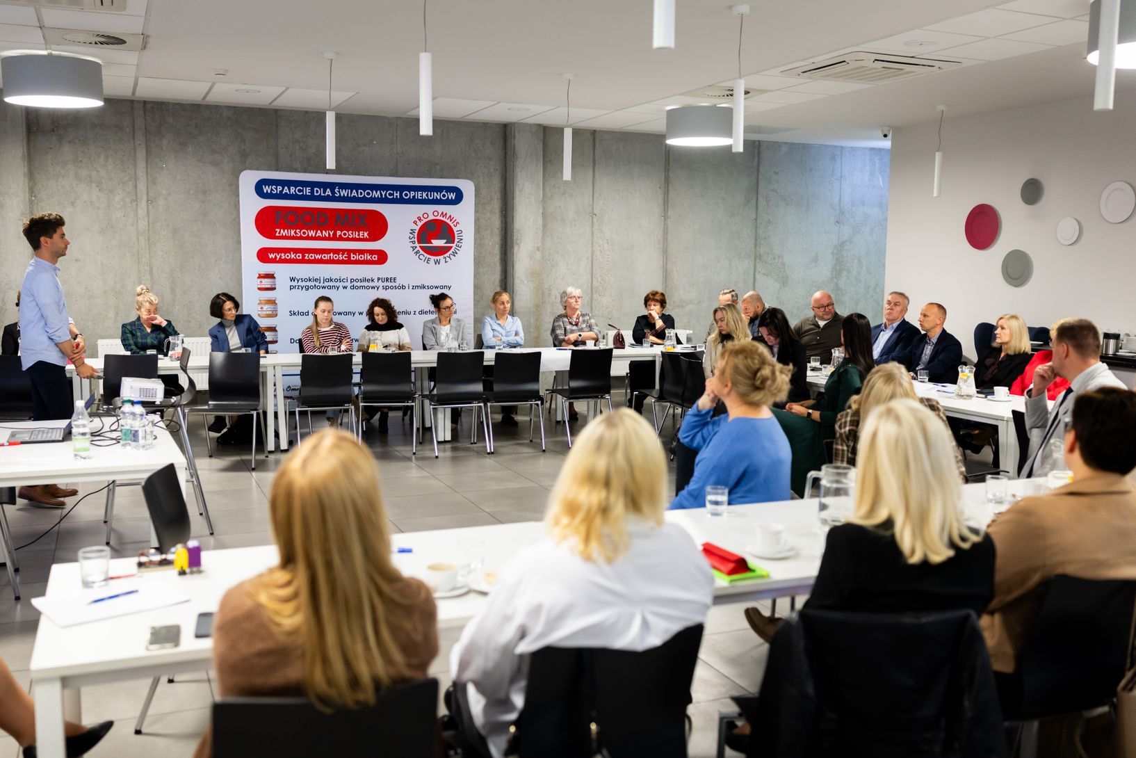 Szeroki plan przedstawia spotkanie grupowe w nowoczesnej sali konferencyjnej. Uczestnicy siedzą przy długich, białych stołach, a jedna grupa zajmuje miejsca na wprost, tyłem do kamery. Młody mężczyzna w jasnej koszuli stoi po lewej stronie, prezentując coś na laptopie. Na ścianie wisi duży baner z tekstem w języku polskim, w tym "WSPARCIE DLA ŚWIADOMYCH OPIEKUNÓW".