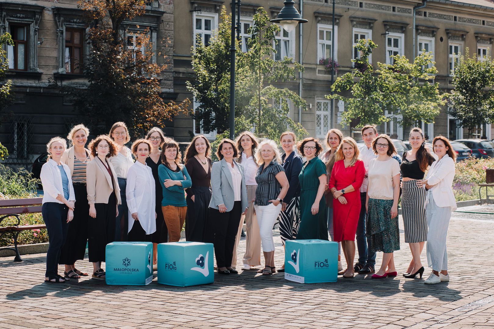 Grupa kilkunastu osób stoi razem na zewnątrz na tle miejskiej zabudowy i zieleni. Uczestnicy są ubrani w styl półformalny i uśmiechają się do zdjęcia. Na pierwszym planie znajdują się turkusowe kostki z logotypami programów Małopolska Lokalnie i FIO Małopolska