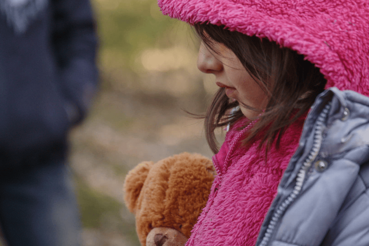 Profil dziewczyznki w rózowej kurtce z kapturem. W tle rozmazany las i sylwetka osoby w czarnej kurtce.