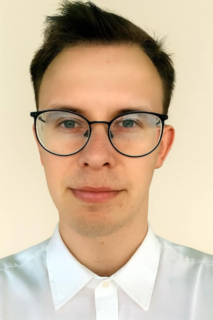 Portretowe zdjęcie młodego mężczyzny w ciemnych oprawkach okularów i białej, eleganckiej koszuli. Mężczyzna ma krótkie, ciemne włosy i patrzy prosto w stronę obiektywu z łagodnym, profesjonalnym wyrazem twarzy. Tło jest jasne i jednolite.