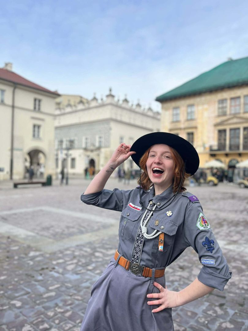 Na zdjęciu znajduje się autorka ogłoszenia w mundurze harcerskim. W tle widać budynki Starego Miasta w Krakowie w okolicy Małego Rynku.