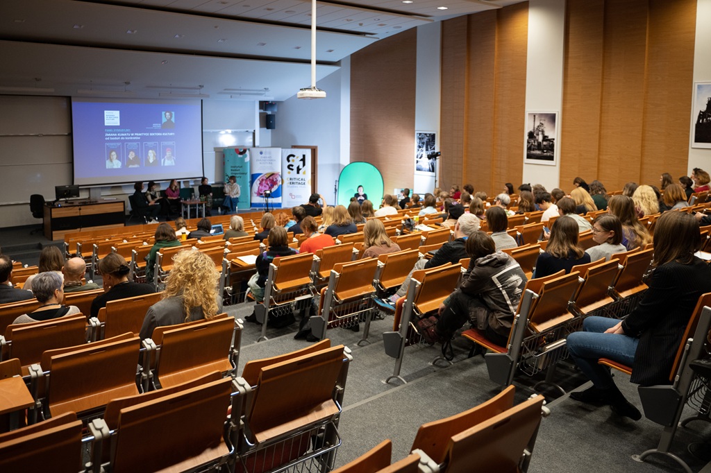 Widok na salę wykładową wypełnioną publicznością siedzącą w rzędach. Na scenie trwa panel dyskusyjny, a prezentacja wyświetlana jest na dużym ekranie. Wnętrze nowoczesne, z drewnianymi wykończeniami i banerami wydarzenia.