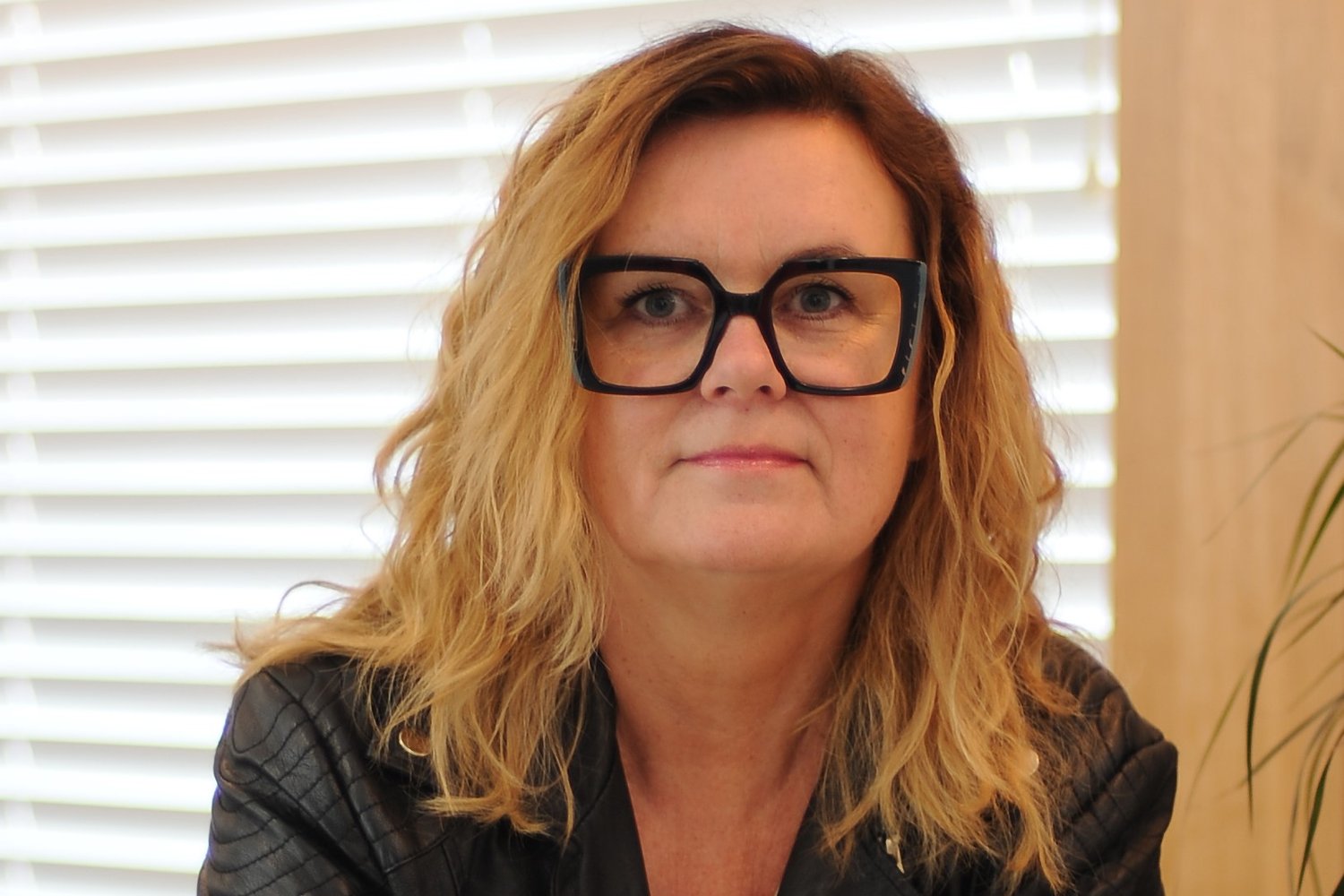Olga Żaryn – kobieta z długimi, falowanymi blond włosami, w dużych czarnych okularach i czarnej skórzanej kurtce, patrzy w obiektyw. W tle jasne żaluzje i zielona roślina.