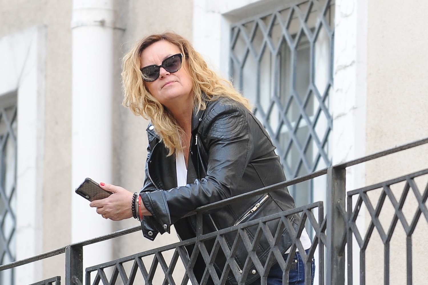 Olga Żaryn w okularach przeciwsłonecznych i czarnej skórzanej kurtce opiera się o metalową balustradę przed budynkiem, trzymając w dłoni telefon.