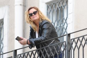 Miniatura Olga Żaryn w okularach przeciwsłonecznych i czarnej skórzanej kurtce opiera się o metalową balustradę przed budynkiem, trzymając w dłoni telefon.