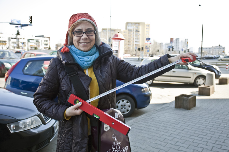 Małgorzata Peretiatkowicz z Niepełnosprawnika stoi na ulicy na Ursynowie. W rękach trzyma poziomicę i miarkę. W tle samochody i bloki mieszkalne